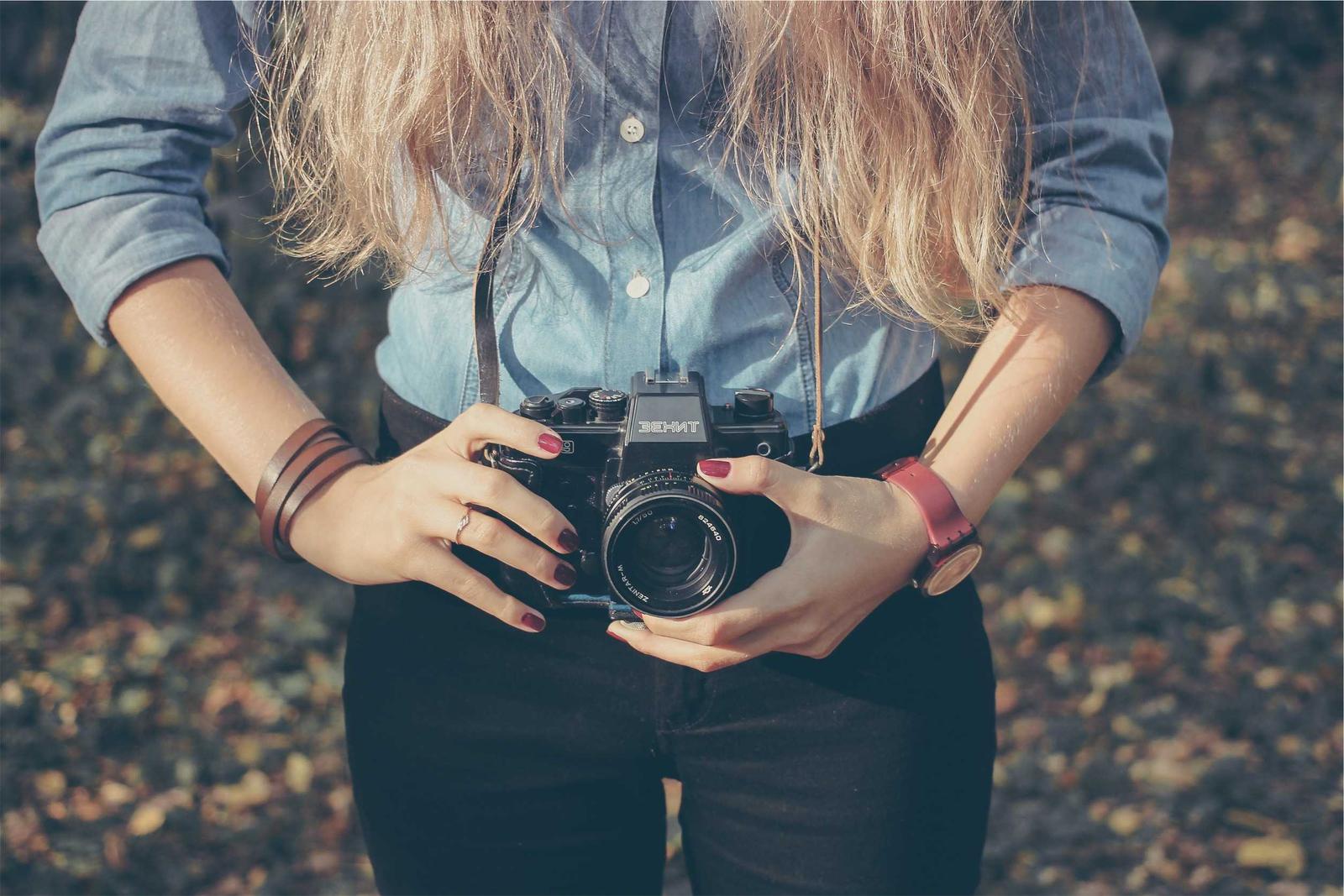 A photographer in a denim shirt holding a film camera at waist height, autumn leaves on the ground behind